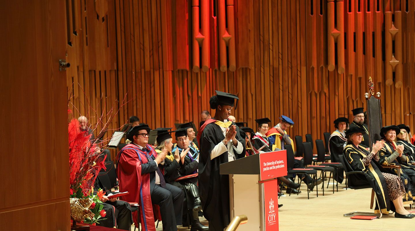 Picture of student speaker Marie Agyare-Wiseborn standing in front of a lectern at the Barbican Centre. She wears a mortarboard and her graduation robe. To her left is a bouquet of  red plants. Behind her is the wooden structure of the Barbican building. She is clapping and looking down at lectern. Behind her, academics are sitting on the stage in their formal academic robes and hats.