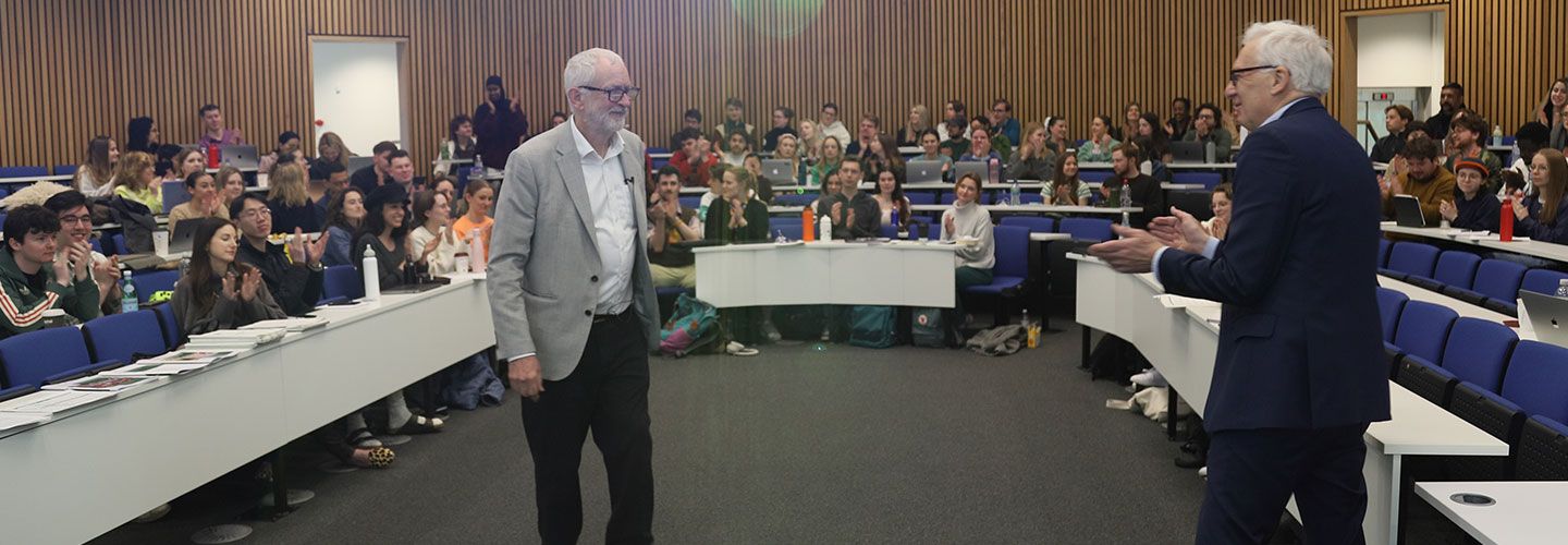 Jeremy Corbyn MP (left) stands and smiles opposite Professor Barney Jones (Lecturer in Journalism) in front of horse-shoe shaped lecture theatre with students smiling and clapping in the backrground