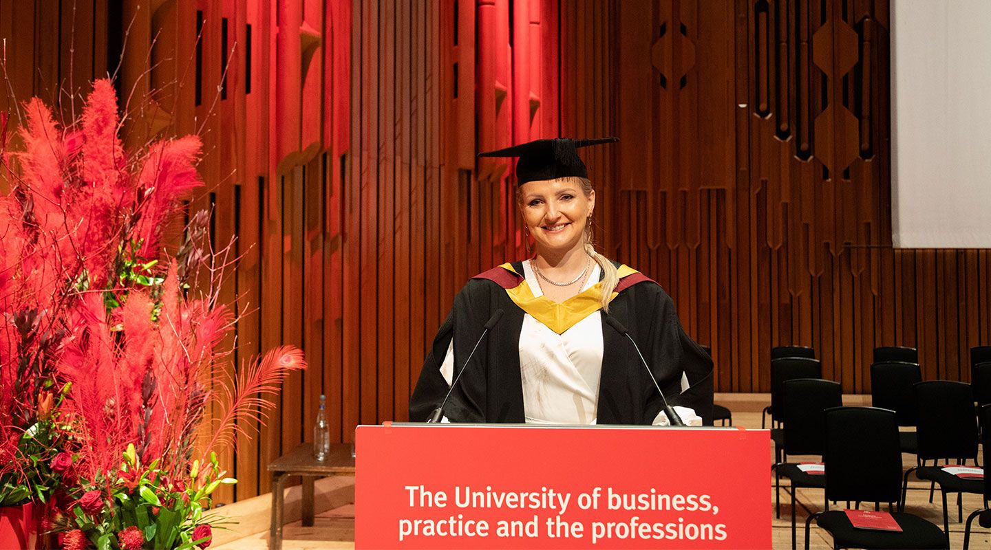 Picture of student speaker Malwina Jaroszewicz standing in front of a lectern at the Barbican Centre. She wears a mortarboard and her graduation robe. To her left is a bouquet of  red plants. Behind her is the wooden structure of the Barbican building. On the lectern is a red sign reading 'University of business, practice and the professions' which is City's slogan