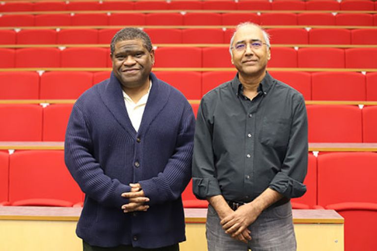 Image of Prof Gary Younge (left) standing next to Prof Inderjeet Parmar (right). They are smiling to camera, standing in front of row of red seats in an amphitheatre.