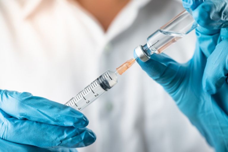 An image of two hands in blue gloves holding a vial of liquid (a vaccine) in one hand and and needle in the other 