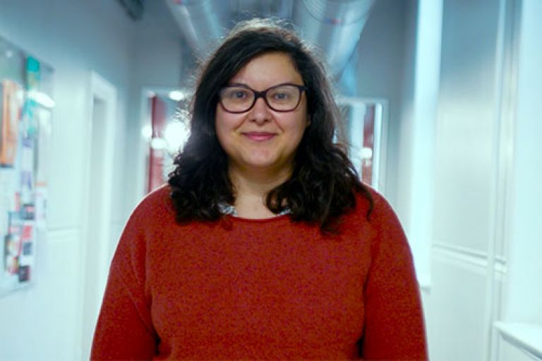 Headshot of Raluca Moise looking towards the camera. She wears glasses and a red a jumper and stands in a corridor on City's campus