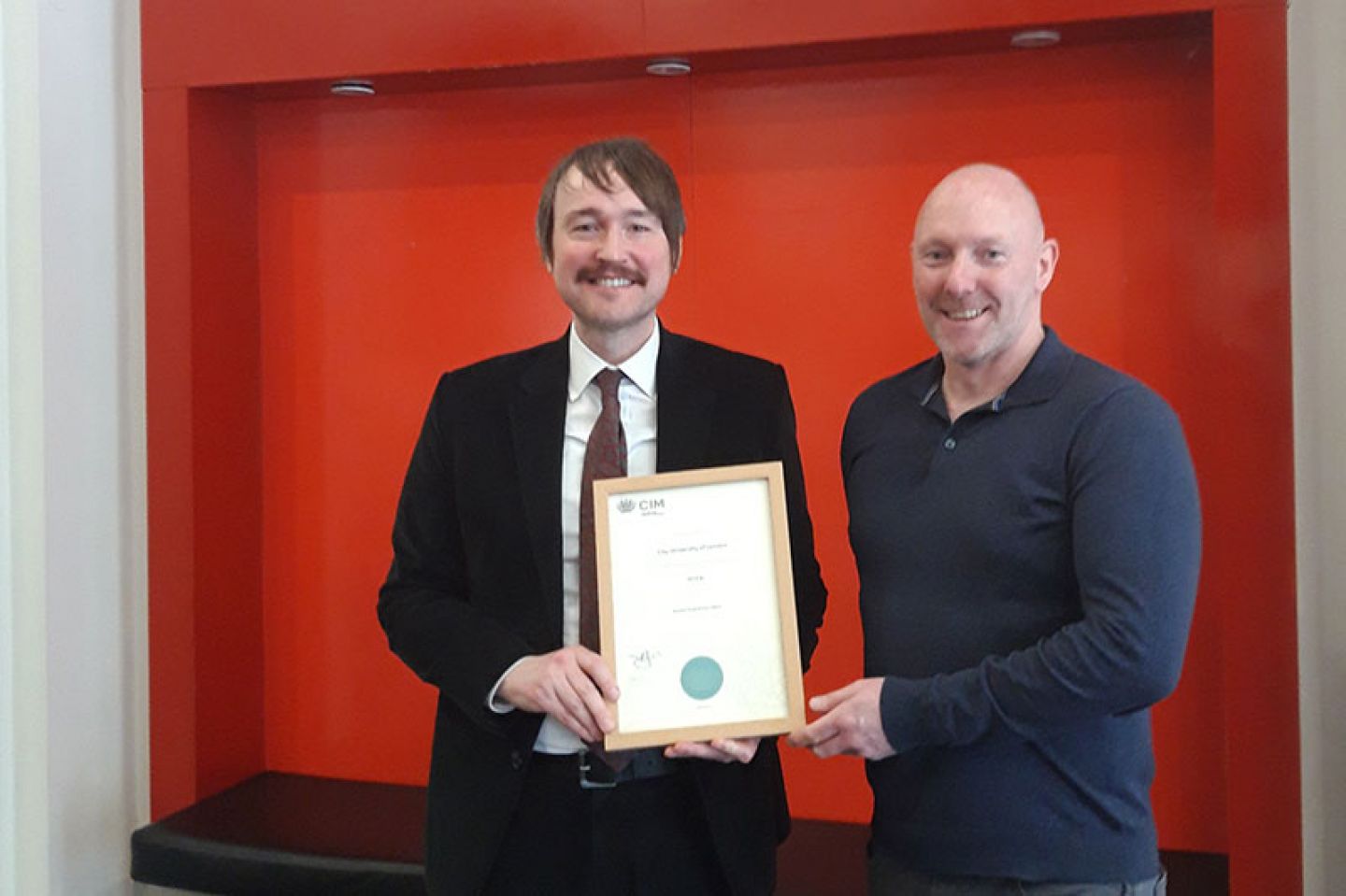 Professor Mattias Frey (left) holds a white certificate handed to him by CIM rep Damian Cuke (right). They stand against a red backrdrop on campus at City