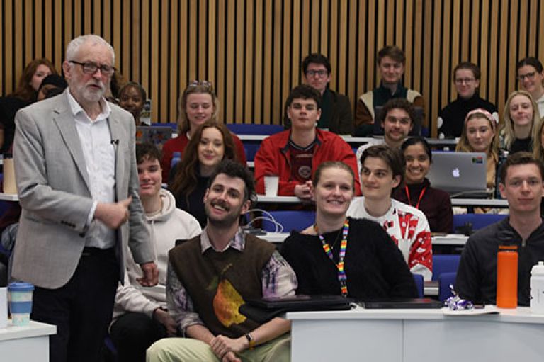 Jeremy Corbyn MP gives a thumbs-up and stands next to journalism master's students who are sitting down on rows of blue chairs in a lecture theatre