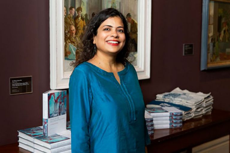 Dr Diya Gupta smiles to camera and stands in front of a desk with copies of her book. On the walls behind her are figurative paintings