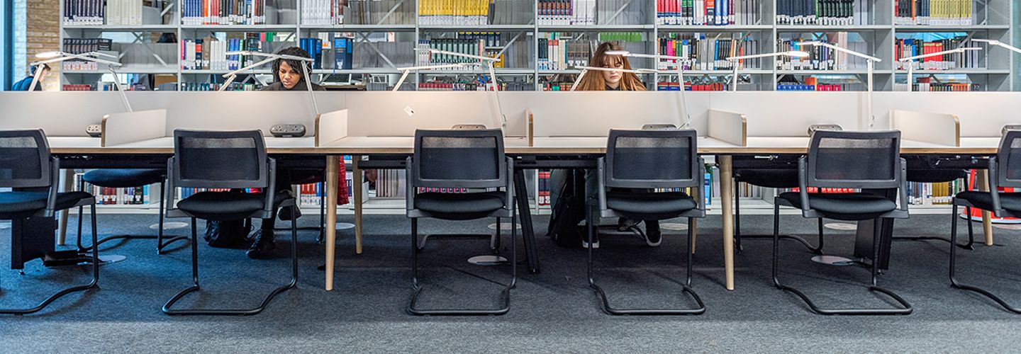 Two females students sat at study desk looking at their laptops with a large bookshelf divider behind
