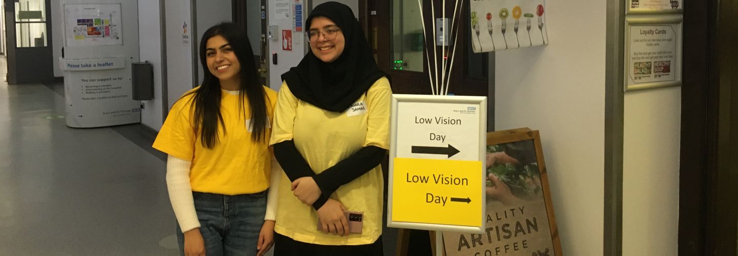 Two second year female optometry students next to a sign pointing to the low vision day event at St Thomas' Hospital