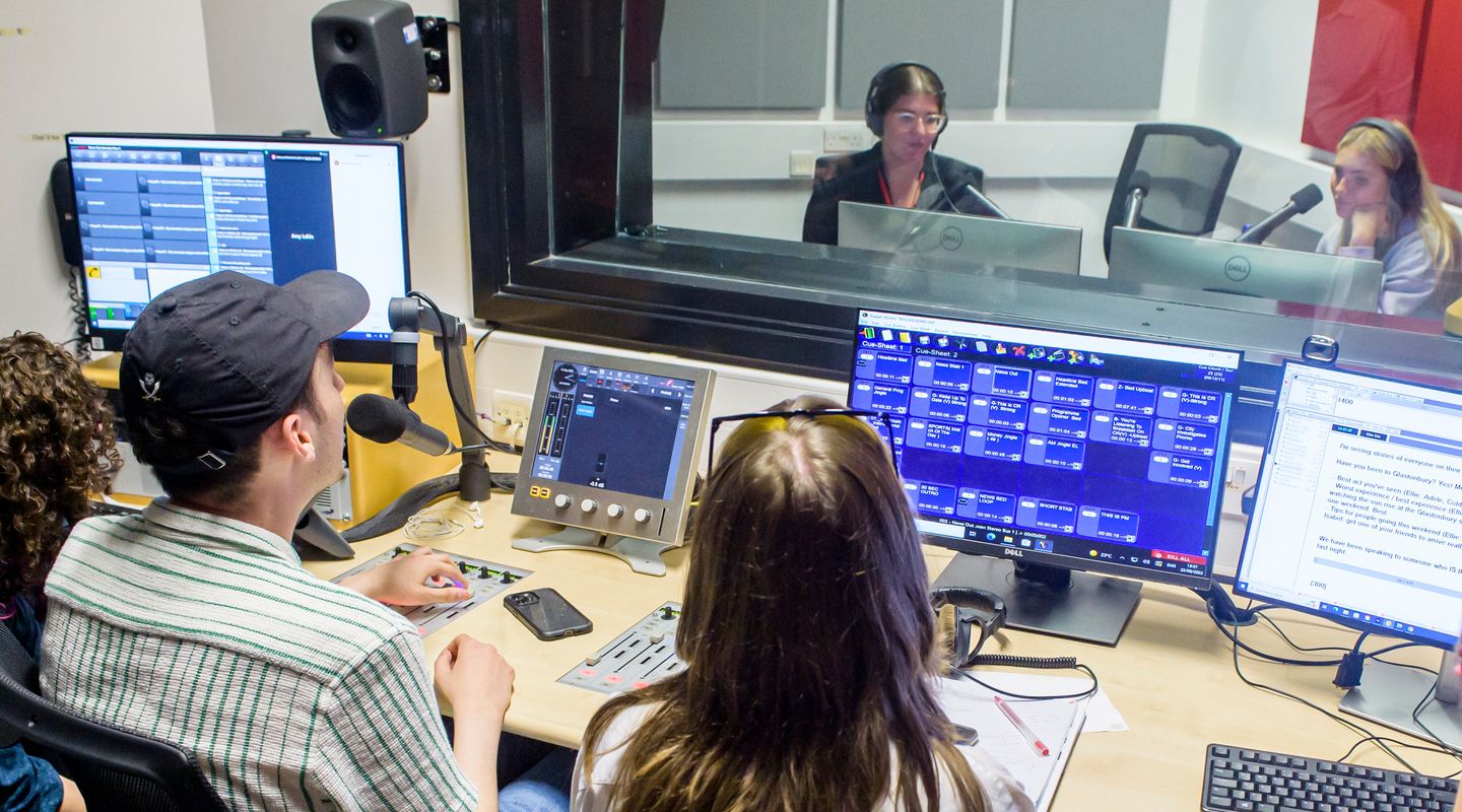A picture of a group of students at the audio studio. On one side, students are in front of computers and use sound mixers. There is a glass separation and on the other side, two other students sit in front of microphones.