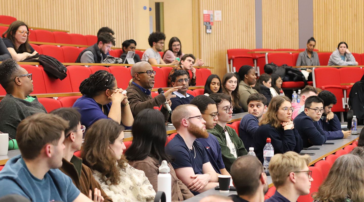 An audience member asks a question. He holds a microhpone and is surrounded by multiple rows of people sitting on red seats in an amphitheatre.