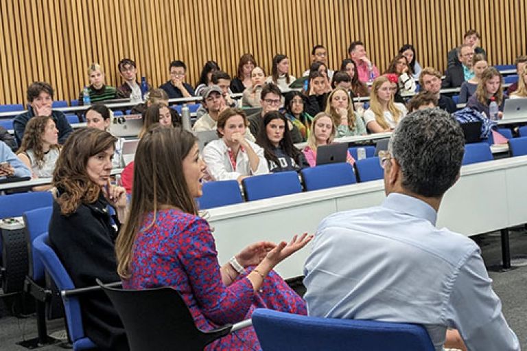 City Question Time panel answers questions. We can see the face of Esme looking at Rosie who speaks, and the back of Kamal facing Rosie. Behind the speakers are journalism students sitting in a horse-shoe lecture theatre at desks, some with laptops in front of them.