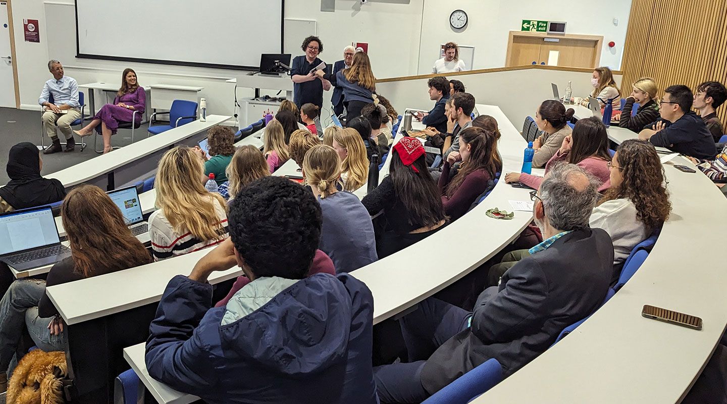 James Ball holds a microphone up to a standing who is standing and asks a question. Around her are rows of students in a lecture theatre with desks in a horse-shoe shape. At the front of the room, speakers Kamal Ahmed and Rosie Wright are sitting on chairs.