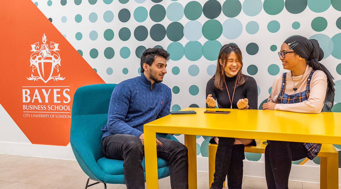 Three students smiling around a yellow table in front of a colourful wall displaying the Bayes Business School logo
