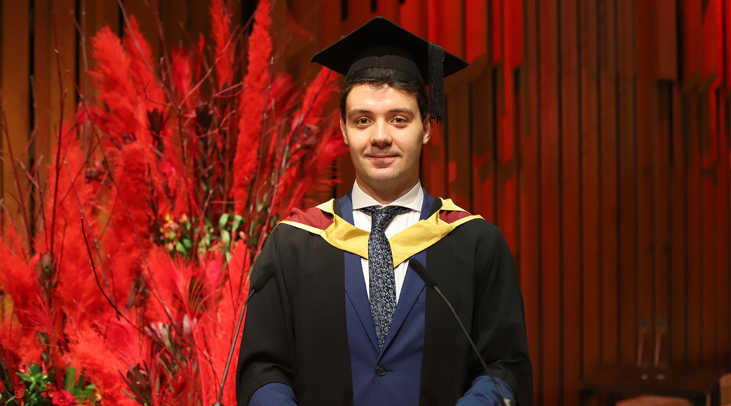 Portrait of student speaker Arturo wearing a black cape and mortarboard in front of a lectern at the Barbican Centre. Behind him is a big bouquet of red plants