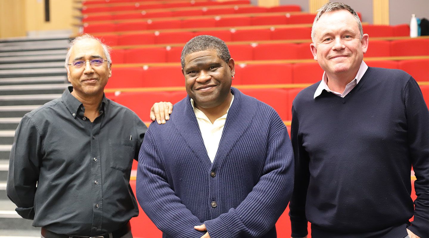 The three men stand and smile to camera. Behind them are rows of red seats in an amphitheatre lecture hall.