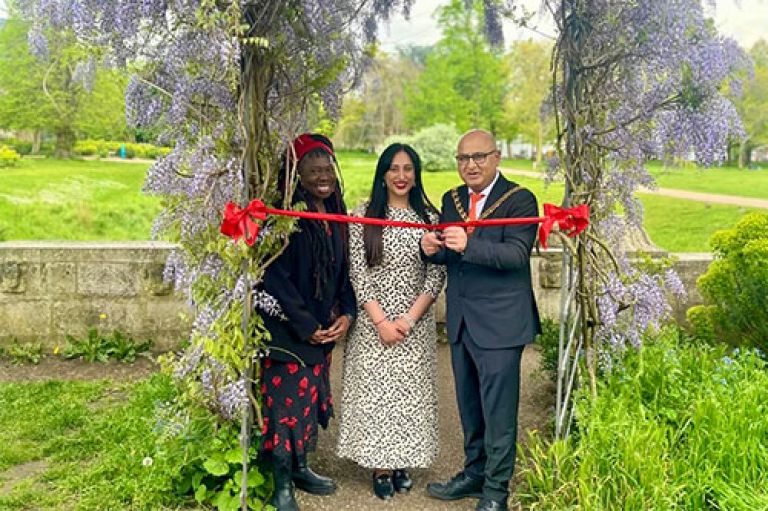 Three people stand under a a wooden arch with wisteria growing on it. At their shoulders is a red sash. The Mayor of Ealing holds scissors to cut this. They stand in a green park.
