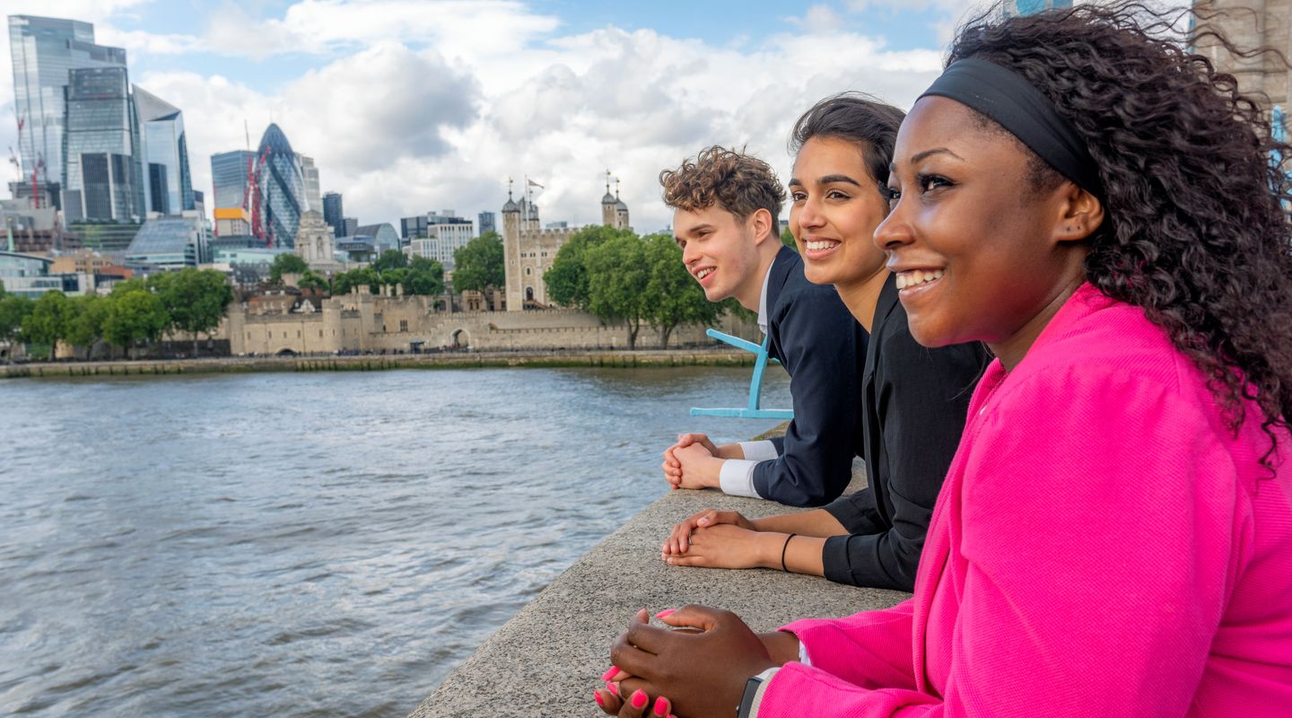 Diverse group of students standing on Tower Bridge, looking over river Thames. Visible behind them is The Tower of London, as well as the City of London.