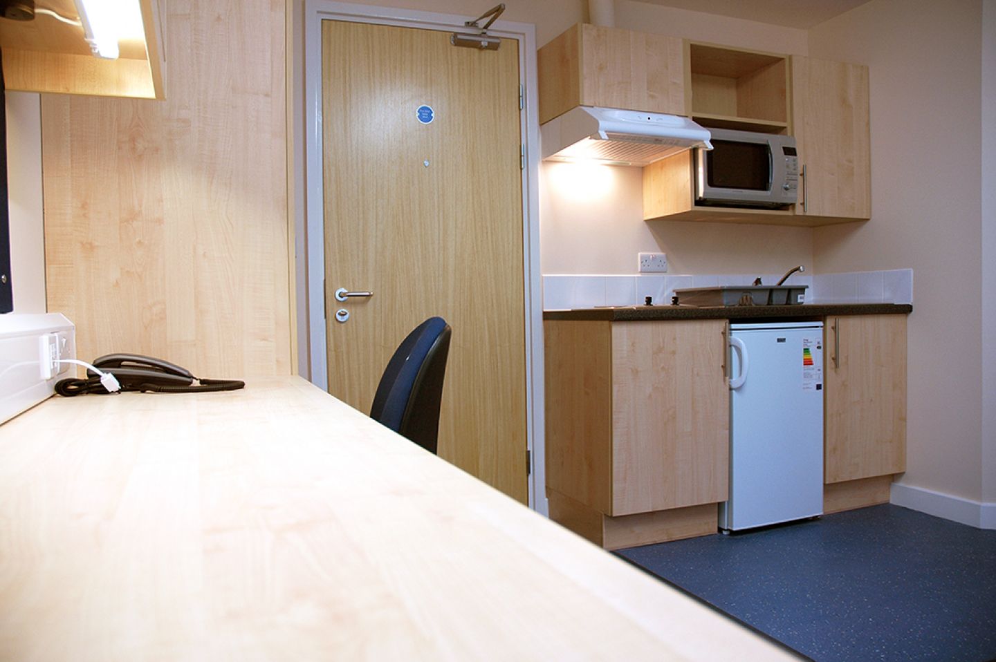 View of a studio room showing the desk, entrance door and kitchen area. The door is a fire door with a closer. The kitchen area has dark blue laminate flooring.