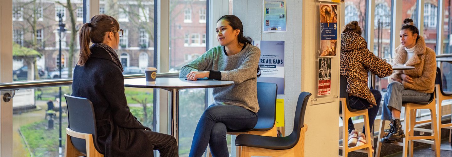 Two pairs of young female students sitting at cafe tables and talking in the communal areas of University Building.