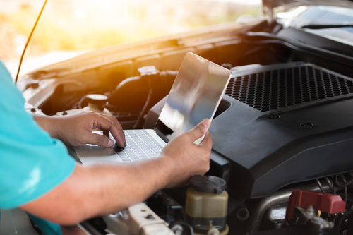 Photo of an automative mechanic using a laptop under the hood of an open car bonnet, near the motor area.